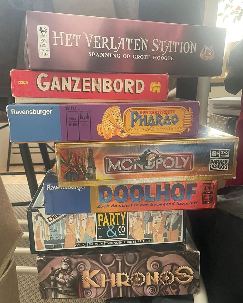 Een stapel bordspellen: Het Verlaten Station, Ganzenbord, Der Zerstreute Pharao, Monopoly, Doolhof, Party & Co en Khronos